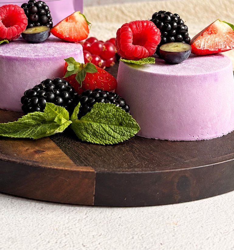 Deux panna cotta violettes à l’ube sur une planche en bois avec framboises, mûres, fraises et menthe